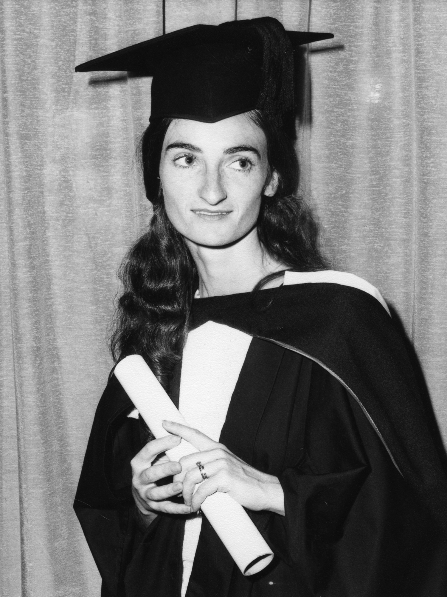 Photograph of an individual with long hair in a graduation cap and gown, holding a rolled up piece of paper in their hand